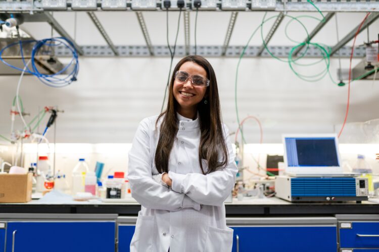 Young woman in a labl coat and safety glasses standing in front of a work bench.