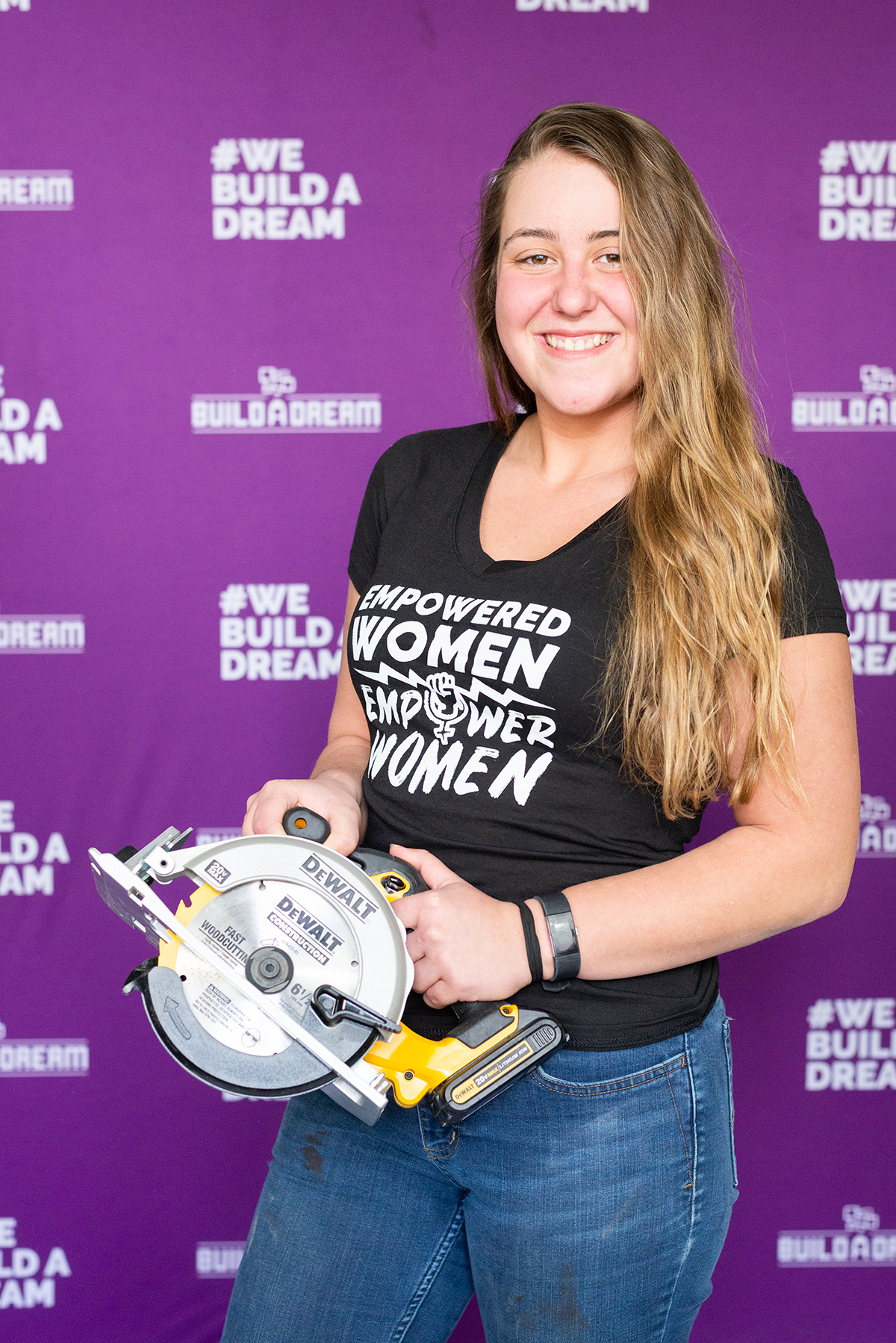 Delaney Krieger stands in front of a purple Build a Dream backdrop and is wearing a black tshirt and holding a circular saw.
